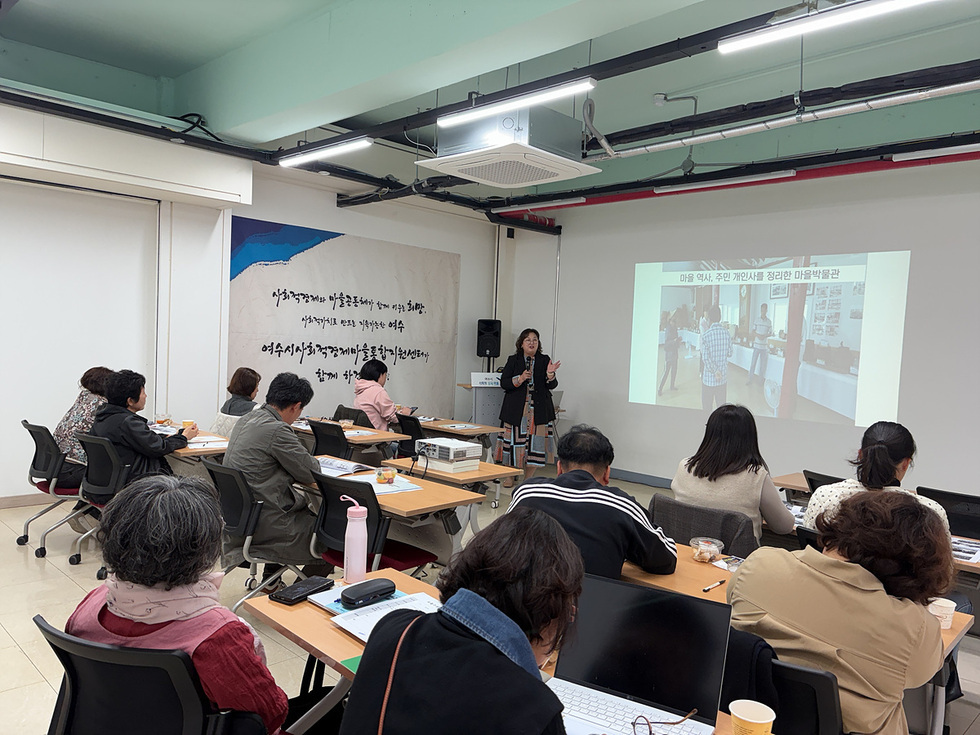 여수시사회적경제마을통합지원센터에서 열린 ‘2025년 마을공동체 입문학교’ 교육 현장. 강사가 마을공동체 사례와 사업계획 작성법을 설명하고 있으며, 7개 마을 주민들이 책상에 앉아 강의 자료를 보며 교육에 참여하고 있다. 스크린에는 마을공동체 활동 사례 이미지가 비춰지고 있다