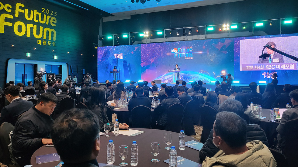 행사장 내부에서 포럼이 진행되는 모습. 원형 테이블에 참석자들이 앉아 있고, 무대에서는 발표자가 연단에서 발언하고 있다. 무대 화면에 ‘KBC 미래포럼’ 문구가 보인다.