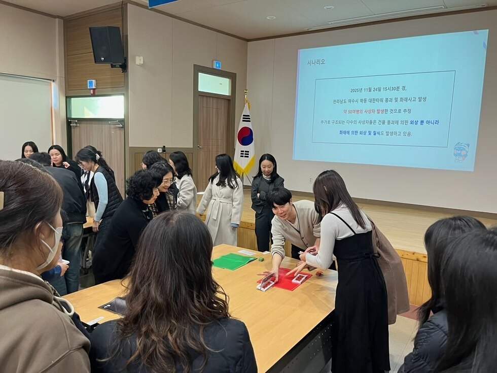 회의실에서 여러 직원들이 책상 주변에 모여 중증도 분류 실습을 진행하는 모습. 한 강사가 책상 위 모형 카드를 배치하며 참가자들에게 설명하고 있다. 뒤쪽 스크린에는 재난 상황 시나리오 안내 문구가 표시돼 있다.