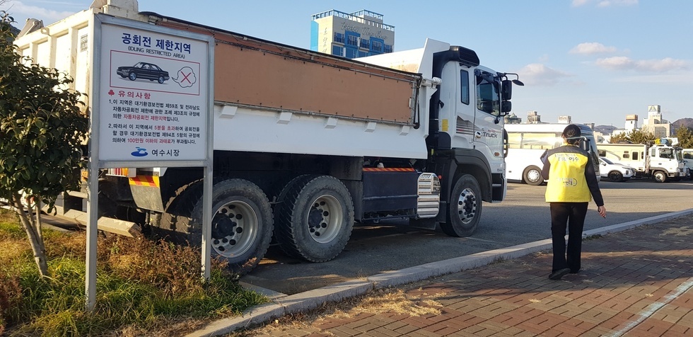 공회전 제한지역 안내표지판 옆에 대형 덤프트럭이 주차된 상태에서 노란 점퍼를 착용한 여수시 단속요원이 현장을 점검하며 걸어가는 모습.
