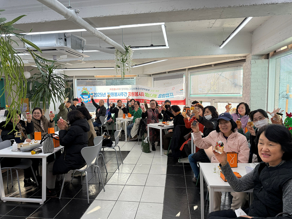 밝은 실내 공간에서 여러 자원봉사자들이 테이블에 둘러앉아 향수 만들기 완성품을 들어 보이며 환하게 웃고 있는 모습. 뒤편 현수막에는 ‘2025년 자원봉사주간 자원봉사자 재능나눔 힐링캠프’,의 문구가 적혀 있다.