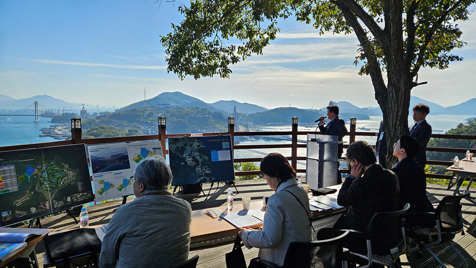 야외 전망 데크에서 바다와 섬이 내려다보이는 풍경을 배경으로 심사가 진행되고 있으며, 참가자들이 자료 패널을 보며 해당내용을 듣고 있다.